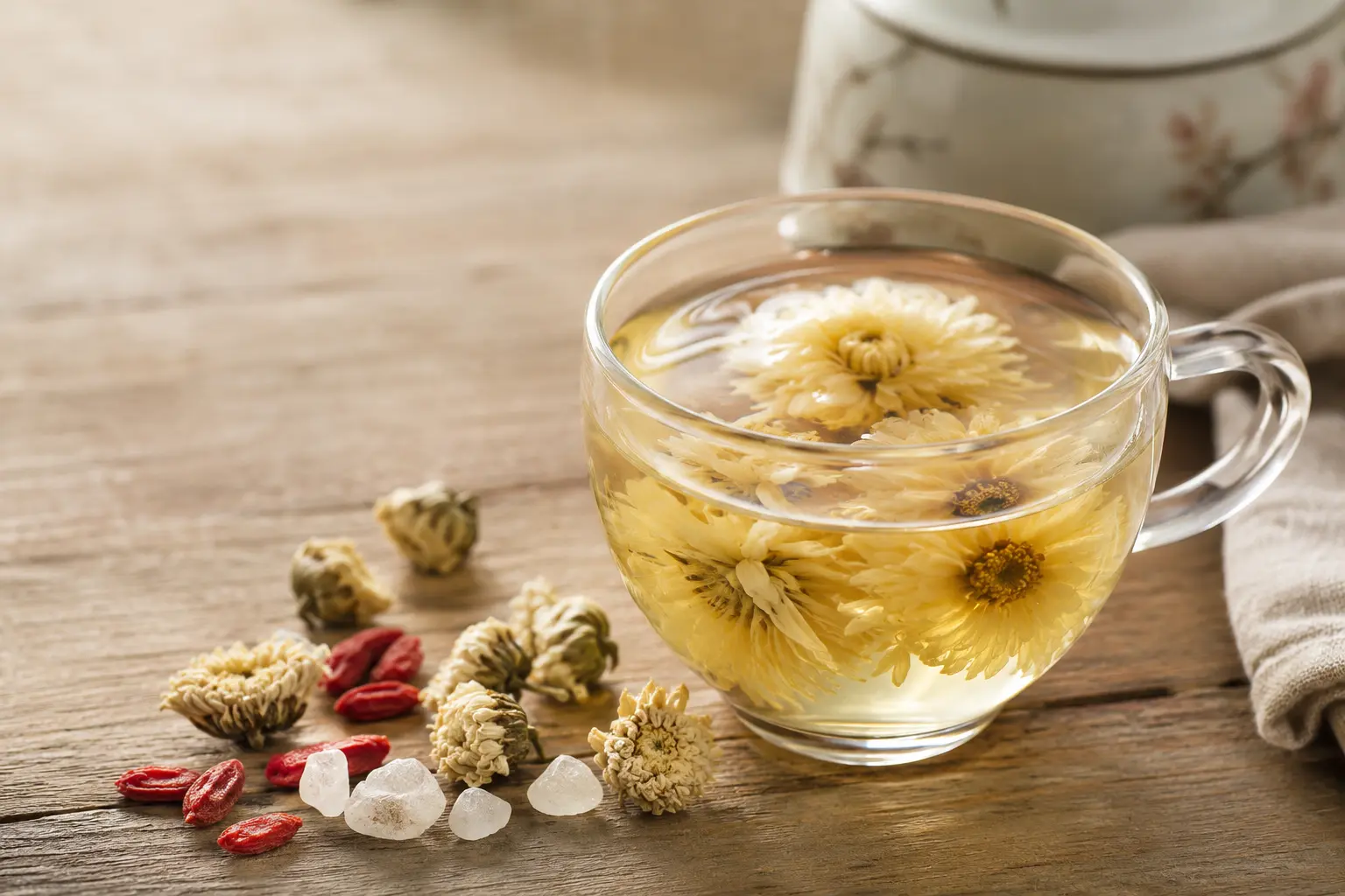 A clear glass of pale golden chrysanthemum tea with whole flowers, dried blossoms and goji berries on a wooden table.